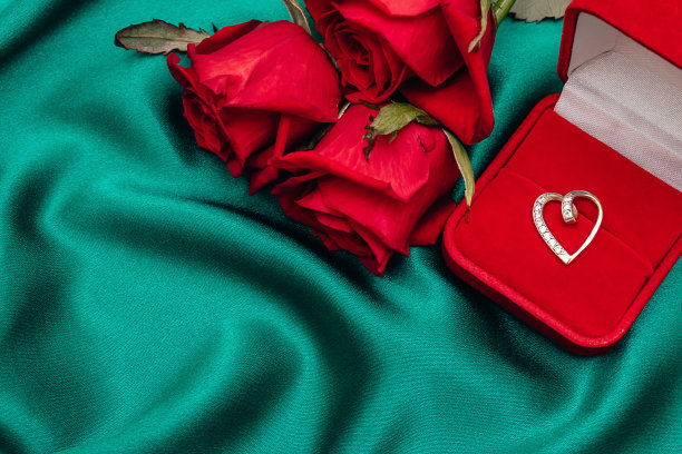 一束红玫瑰和一个红色天鹅绒礼盒，钻石心形吊坠在绿色丝绸背景。情人节的概念。复制空间的文本，选择焦点。图片下载