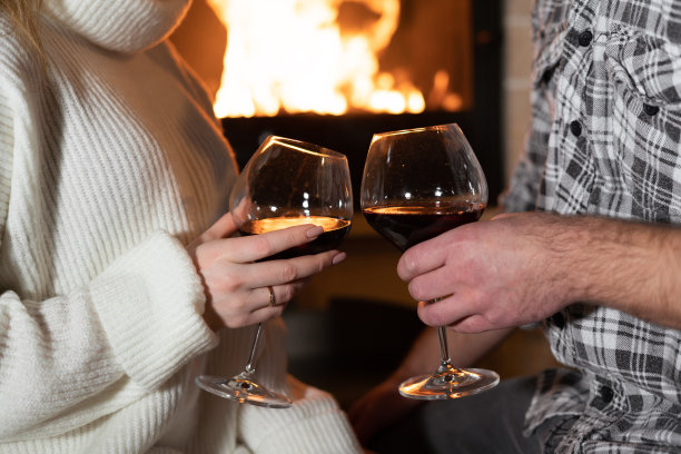 在一个假日的晚上，一个男人和一个女人站在壁炉旁，他们的手握着盛着红酒的玻璃杯。家的舒适和爱的氛围。图片下载
