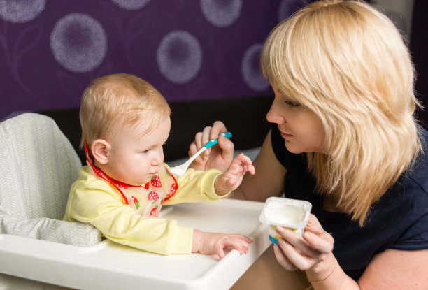 妈妈用罐子喂婴儿喝酸奶。那孩子正坐在一张高椅子上。一条围裙挂在胸前图片下载