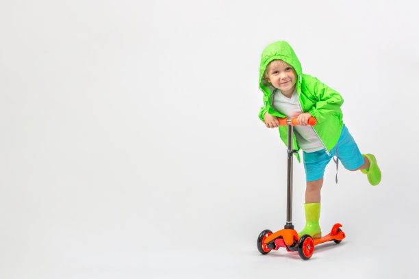 欢快的孩子孩子在明亮的绿色夹克兜帽和橡胶靴子骑摩托车在白色的背景与拷贝空间。摄影棚里一个4岁的男孩。童年和活跃的休闲概念。图片下载