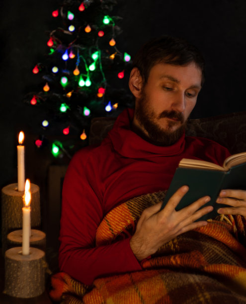 一名身穿红色毛衣的大胡子男子在燃烧着的蜡烛和装饰着节日气氛的圣诞树旁看书。图片下载