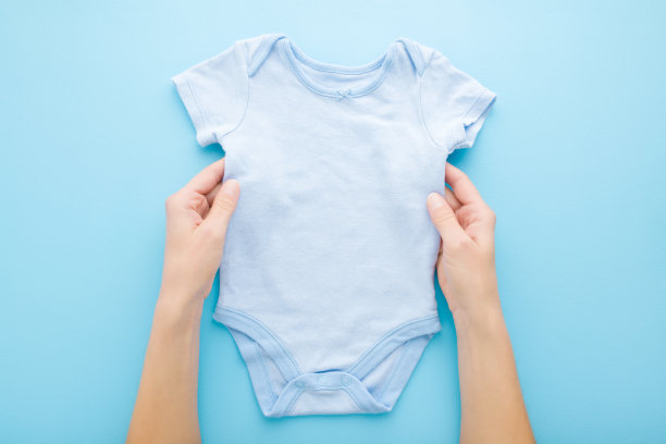 年轻的成年母亲的手抱着新的婴儿男孩紧身衣在浅蓝色的桌子背景。柔和的颜色。特写镜头。视角拍摄。为服装上的文字或标识留出空位。自顶向下的观点。图片下载