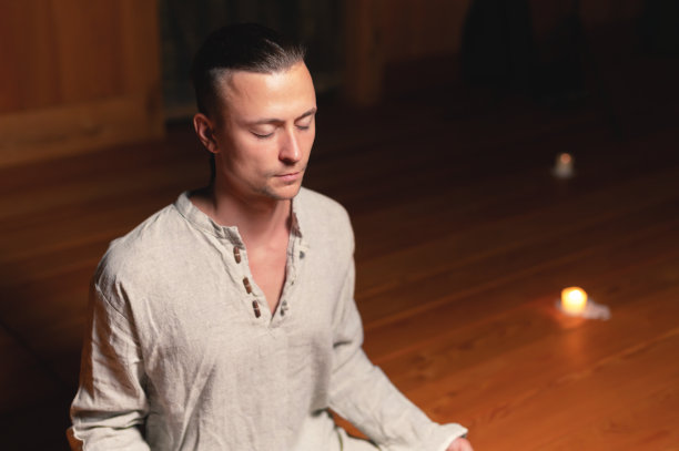 在一间昏暗的木屋子里，一名身穿修身和冥想服的男子以莲花的姿势坐着，手持红色念珠来集中注意力图片下载