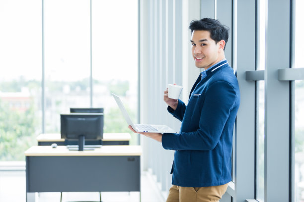 快乐心情亚洲年轻商人工作手提电脑和拿着咖啡杯在窗口在办公室的背景。图片下载