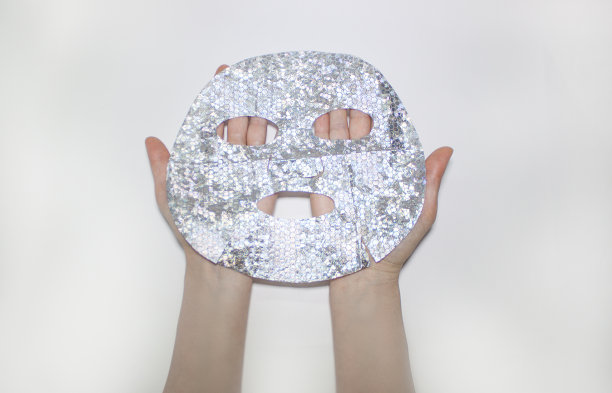 女性手里拿着一个布料全息银色面膜。在白色的背景上。从以上观点。美容、医药和保健。图片下载