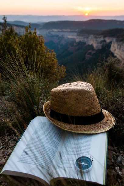 旅行的概念。草帽与旅游冒险书在山上以日落或日出为背景。故事，想象，脱离常规。图片下载