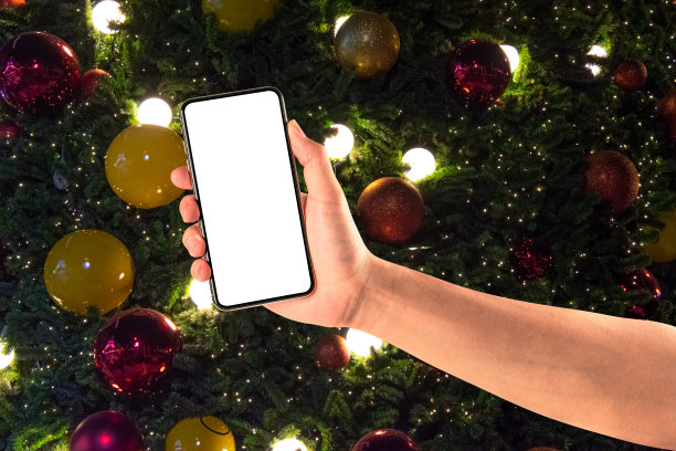 近距离手持手机孤立在白色的背景与灯光装饰圣诞树挂着金黄色和红色的球。图片下载