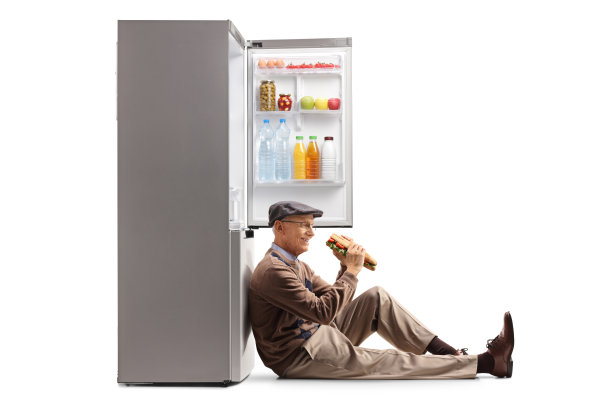老人吃着三明治，坐在地板上，靠在一个敞开的冰箱上图片下载