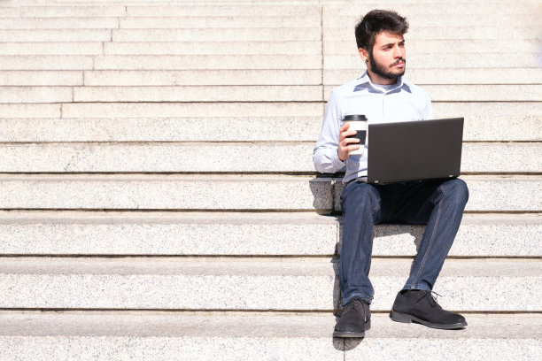 一个年轻的商人坐在楼梯上，用他的笔记本电脑拿着一个咖啡杯。城市街道上的商人。图片下载