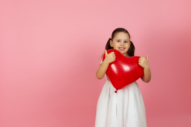活泼的小女孩穿着白色连衣裙，拿着心形的红色气球，在两只手上竖起大拇指，孤立在粉红色的背景上图片下载
