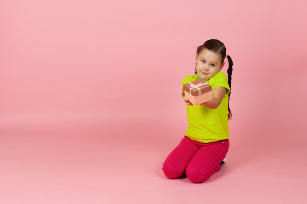 一个梳着马尾辫、穿着淡绿色t恤和红色裤子的假女孩递给我们一个礼品盒，盒子放在地板上，在粉红色的背景下显得孤立图片下载
