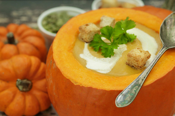 图片中，橙色南瓜的盖子被打开，里面装满了自制的南瓜汤，酸奶和欧芹装饰，金属勺子，一碗一碗的南瓜籽和油炸面包丁，新鲜采摘的葫芦，木质背景，从高处看，重点放在前景图片下载