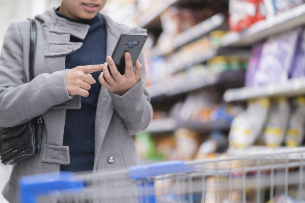 特写亚洲女性手持智能手机在超市里用手推车购物车和一排排产品比价图片下载