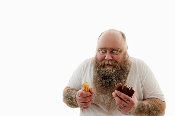 白色背景上的一幅孤立的肖像，一个肥胖的大胡子男人，手臂上有纹身，手里拿着巧克力棒、蛋糕或泡芙，贪婪地看着它们图片下载