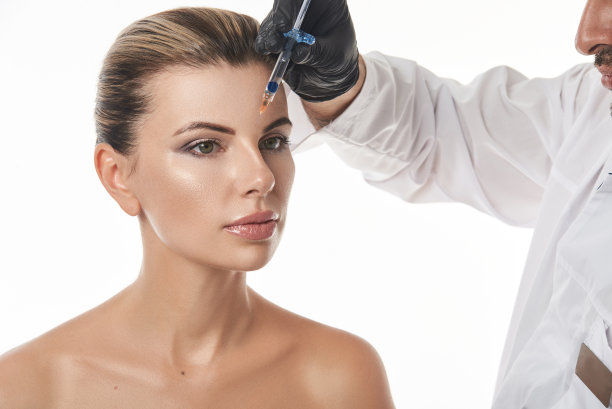 美丽的女人的脸和医生的美容师或整形外科医生的手用注射器使化妆品注射在前额。注射美容图片下载