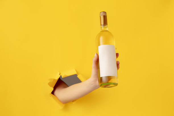 女性手拿一瓶白葡萄酒穿过黄色的纸背景。图片下载