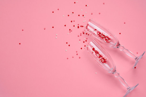 两个香槟酒杯，粉红色的背景上撒满了红心形状的五彩纸屑。情人节的概念。俯视图，复制空间。图片下载