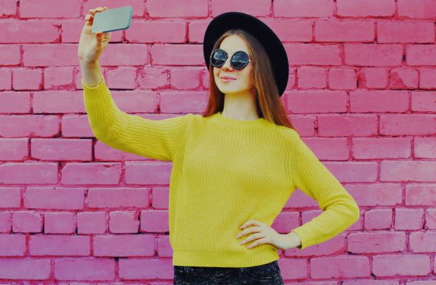 一名微笑的年轻女子在粉色砖墙背景下用智能手机自拍图片下载