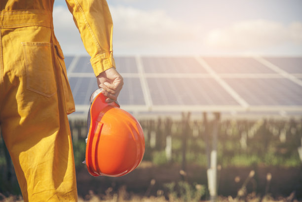 清洁和可持续能源概念。建筑工程师或电气工人拿着橙色安全帽在太阳能电池板的背景下工作。工头穿着安全服，看着发电厂。图片下载