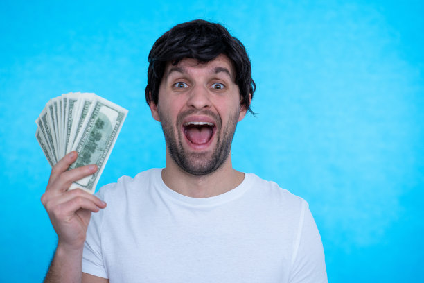 银行储蓄。一个年轻英俊自信的男人手里拿着美元，开心地看着蓝色背景下的相机图片下载