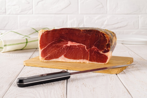 1公斤重的火腿片和锋利的长刃刀放在切菜板上，放在白色的木桌上。美味的西班牙塞拉诺火腿。传统腊肉美味。图片下载