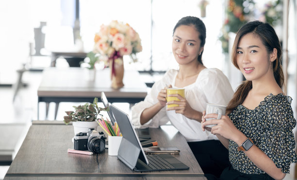 这是两个年轻的大学生拿着杯子，微笑着看着镜头，坐在共同工作空间的桌子上。图片下载