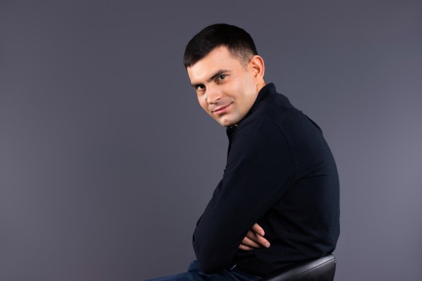 一名穿着黑色衬衫、坐在真皮椅子上、灰色背景的时尚男子看着摄像机。档案照片。图片下载