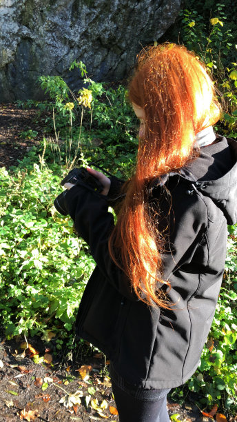 红发少女学习摄影的图像，用数码单反相机拍照，聚焦镜头在郊外，穿着黑色防雨外套，看着相机屏幕的取景器图片下载