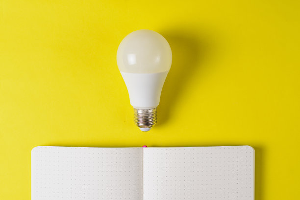 打开一个白色笔记本，明亮的黄色背景上有一个灯泡。灵感搜索概念的个人笔记，生活规划和业务。思想的起源。文本的空闲空间。图片下载