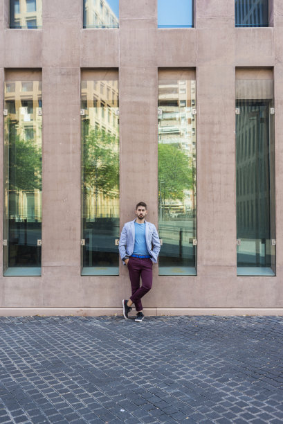 年轻的大胡子男子，时尚模特，在城市背景穿着休闲服装，倚在一幢办公楼的墙上图片下载
