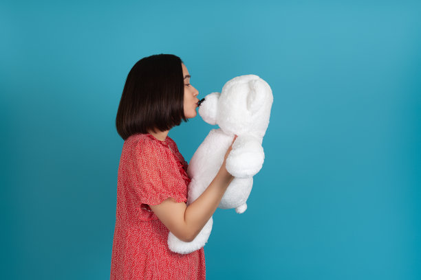 一名身穿红色连衣裙的亚洲女子侧身对着镜头，亲吻蓝色背景下一只白色泰迪熊的鼻子图片下载