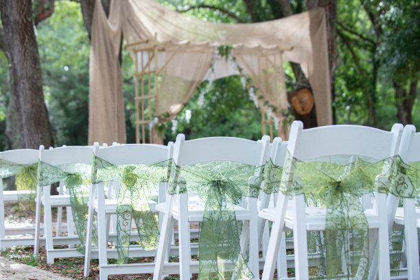 在波西米亚户外婚礼仪式上，配有绿色蕾丝蝴蝶结和饰带的白色折叠椅排列在棕色布制祭坛前图片下载