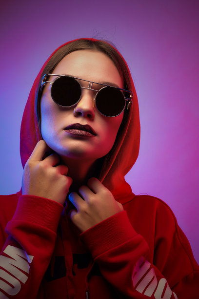 女孩在时尚的霓虹灯下在工作室摆姿势。女人在圆形太阳镜和帽衫与色彩斑斓的光效果。图片下载