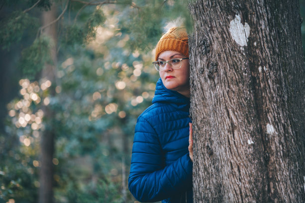 一个漂亮的女人戴着一顶冬天的帽子从树林里的一棵树后面探出头来图片下载