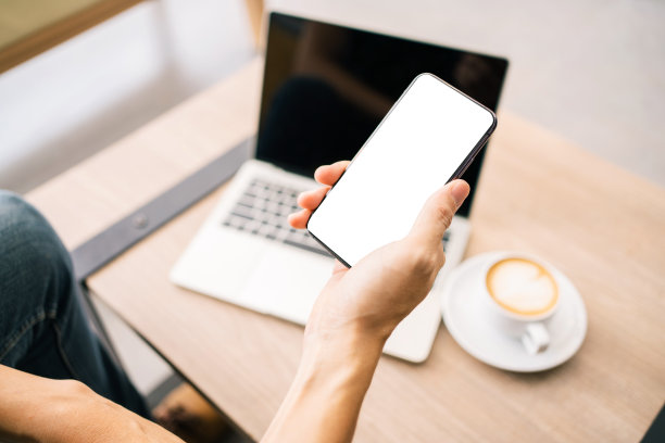 手机模型图像与空白白屏幕。男性手握手机发短信在桌子上，笔记本电脑和咖啡杯放在桌子上的背景。空白的广告文字。图片下载