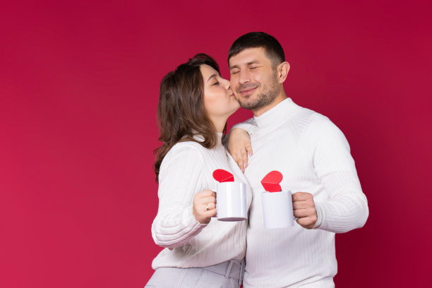 女人在红色背景上亲吻心爱的男人。年轻人拿着白色的茶杯。图片下载