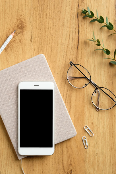 当代办公桌俯视图。平板手机与空白屏幕模型，纸笔记本，眼镜，女性眼镜，桉树叶在木制背景。现代的自由职业者的工作空间。图片下载