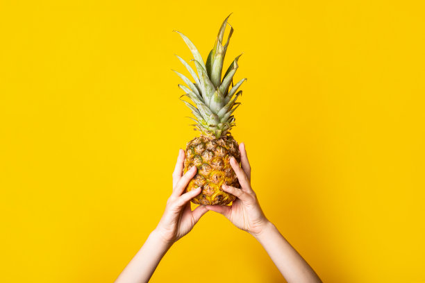 女性的手拿着一个完整的成熟的菠萝，背景是撕破的黄色。图片下载