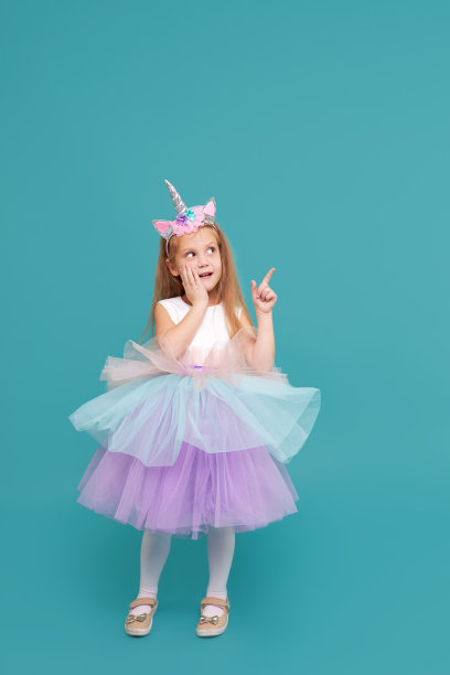 独角兽女孩。有趣的脸。可爱的小女孩在优雅的薄纱裙和独角兽头带庆祝生日派对在彩色的背景。指向右边图片下载