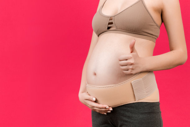 特写孕妇显示拇指向上和穿着弹性怀孕绷带，使疼痛消失在粉红色的背景与复制空间。骨科腹部支撑带概念图片下载