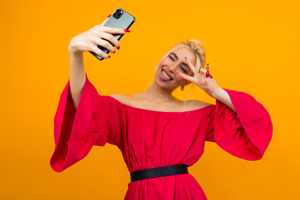 一个年轻的微笑的女人在一个黄色的工作室背景的手机上做自己的照片优雅的红色连衣裙图片下载