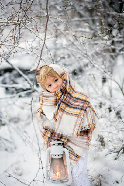 金发碧眼的蹒跚学步的小男孩，穿着手工织的白色外套，在雪地里举着灯笼图片下载
