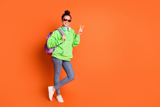 照片迷人的年轻女士穿着绿色运动衫背包眼镜显示v标志孤立明亮的橙色背景图片下载