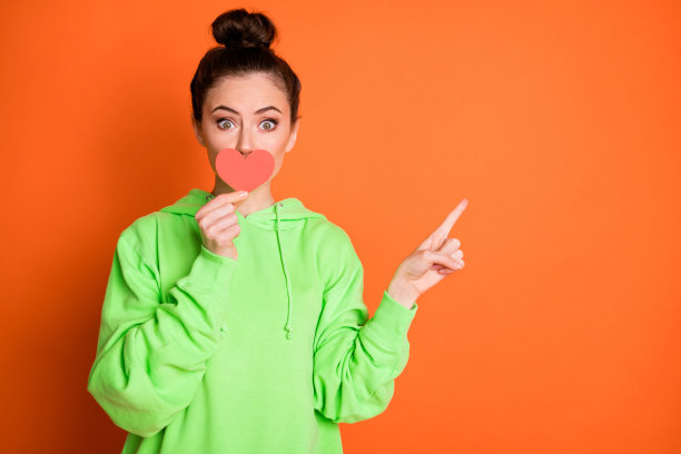 照片肖像可爱的女孩覆盖嘴与小红心卡片指向的手指空白孤立在生动的橙色背景图片下载