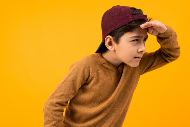 穿着棕色毛衣和棒球帽的欧洲帅哥正在寻找黄色背景上的目标图片下载