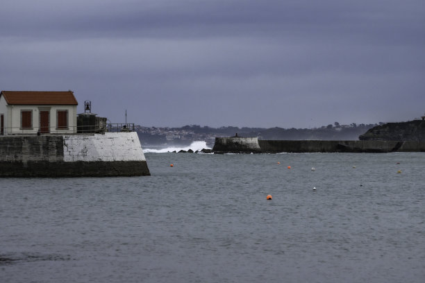 这是法国巴斯克地区索卡港木板路的日出照片，你可以看到白天的第一缕阳光，港口的入口，海浪拍打着海面上的岩石。前景是一幢红屋顶的白色房子。图片下载