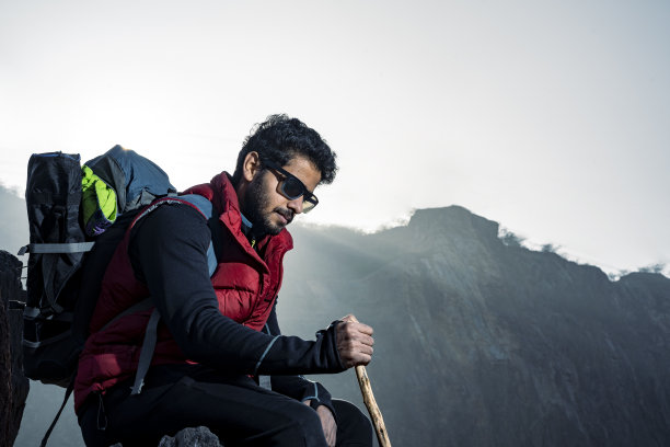 一位年轻的印度登山家背着背包，拄着一根拐杖，坐在山顶的悬崖上，欣赏着山顶上的美景，这张照片拍摄于日落时分。图片下载