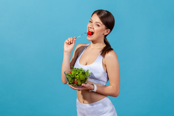 快乐的女人穿着白色运动服，身材苗条，吃着大碗里鲜红多汁的西红柿，配上新鲜的沙拉，素菜午餐，健康饮食，节食图片下载