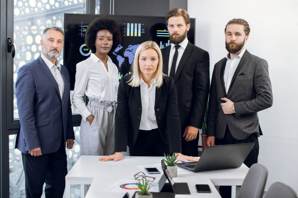 五名不同种族的商务人士组成的团队，前景是一位成熟的金发职业女性商务领袖，站在办公桌前，带着笔记本电脑，在现代的办公室里图片下载