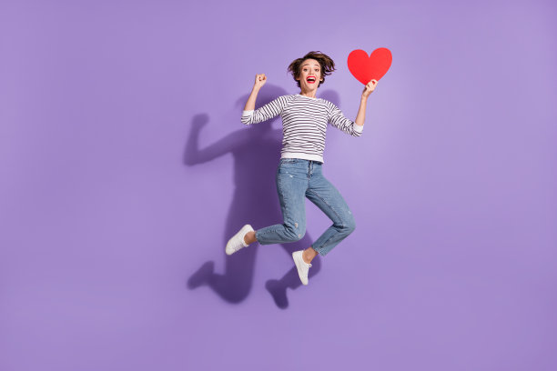 全身照片可爱的年轻女子穿着休闲装跳抱着看红纸心孤立的紫罗兰色背景图片下载